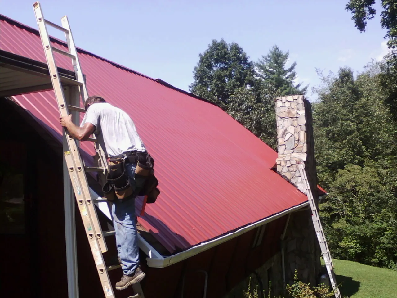 Roofer on a ladder beside a red metal roof and stone chimney — Soffit & Fascia Repair by Roofing Brothers in Tazewell